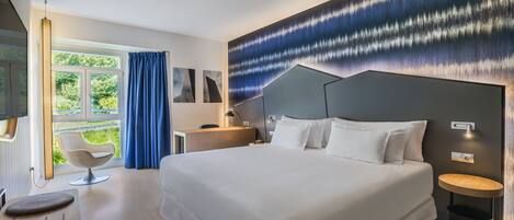 A modern hotel room with a large bed, a desk, a chair, and a view of greenery outside.