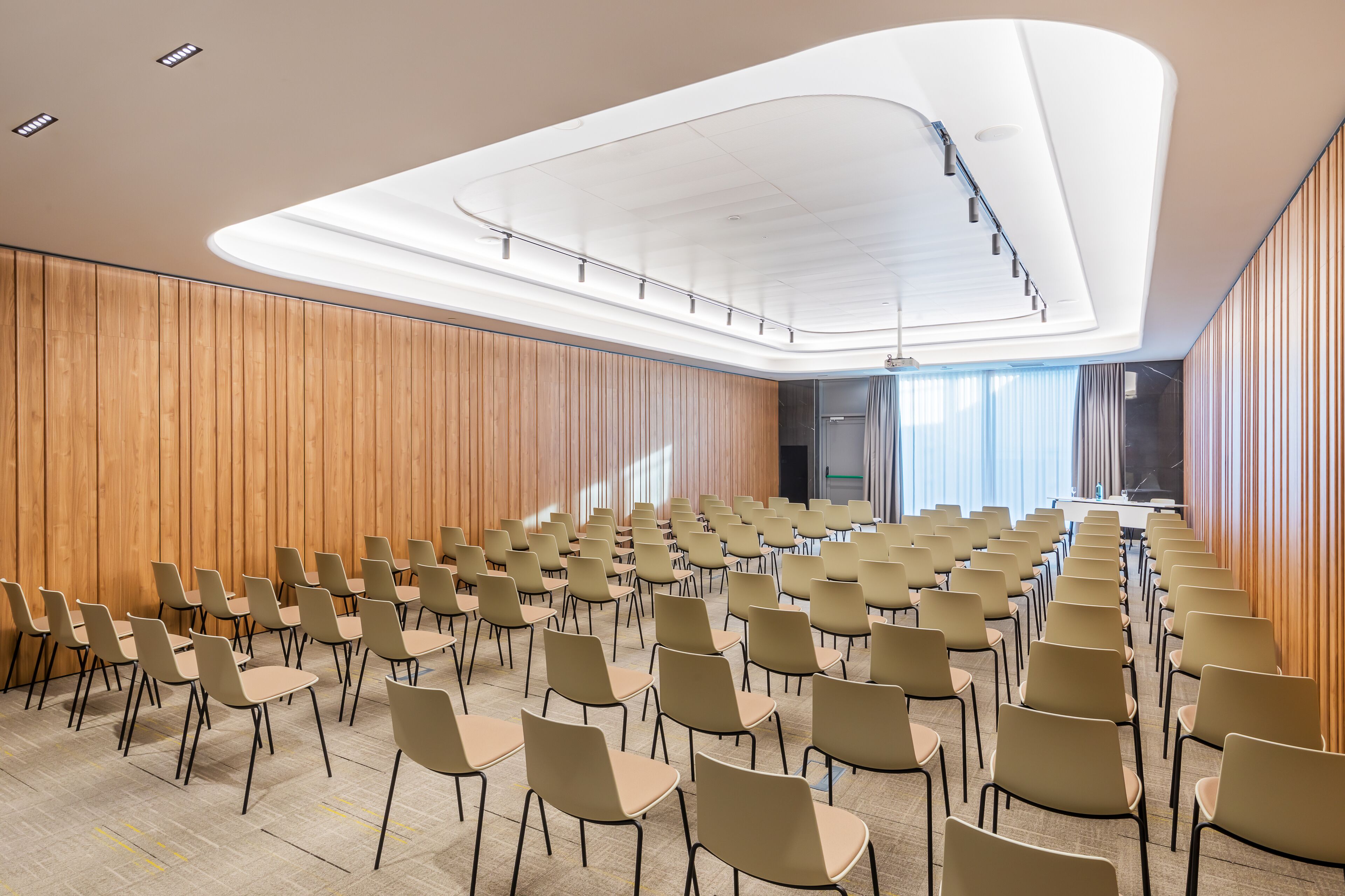 Une salle de conférence avec des rangées de chaises, un mur recouvert de panneaux en bois et un design moderne au niveau du plafond.