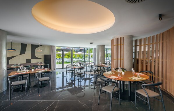 Un comedor moderno con mesas y sillas redondas, un elemento de techo iluminado y vistas al exterior.