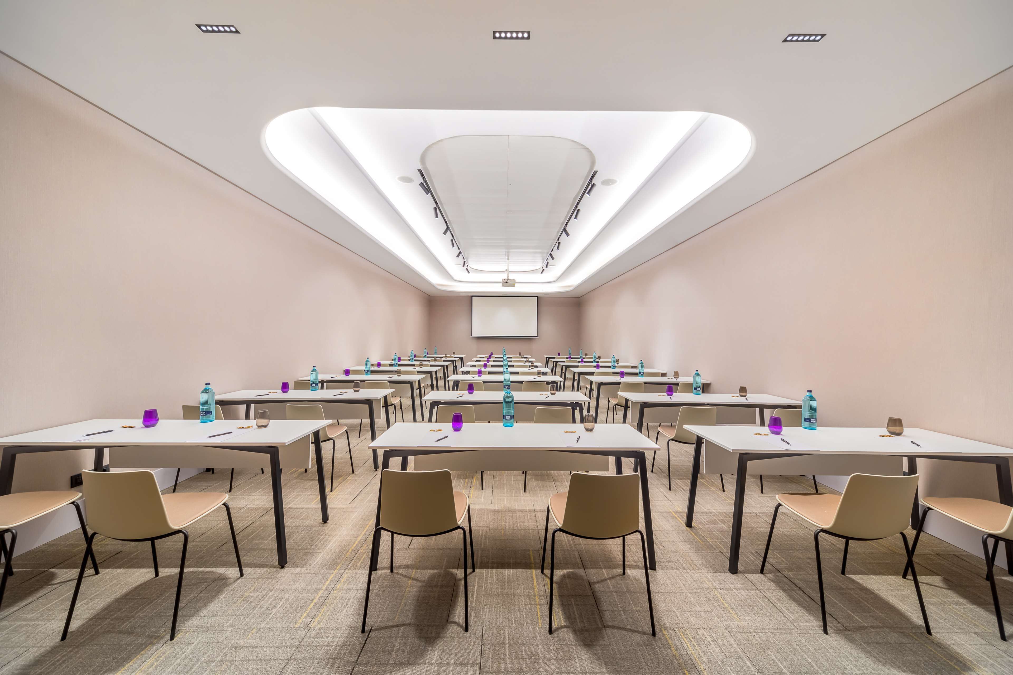 A conference room with multiple tables and chairs, equipped with water bottles and glasses, and a large screen at the front.