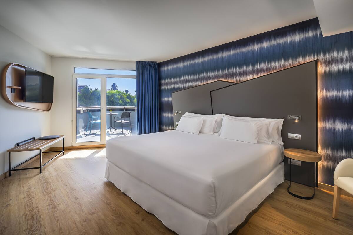 A modern hotel room with a large bed, a TV mounted on the wall, and a balcony with a table and chairs.