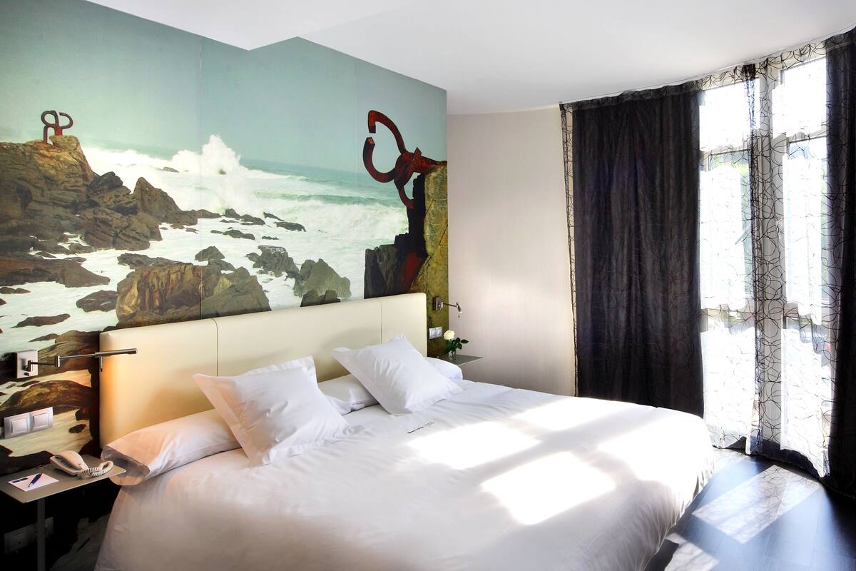 A hotel room with a large bed, a painting of a coastal scene, a bedside telephone, and a window with curtains.