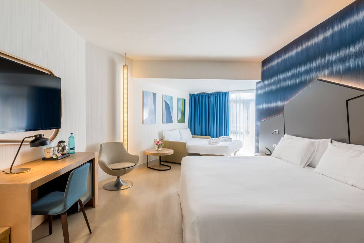 A modern hotel room with a large bed, a desk with a computer, a chair, a small table with a vase, and a TV.
