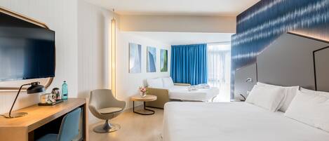 A modern hotel room with a large bed, a desk with a computer, a chair, a small table with a vase, and a TV.