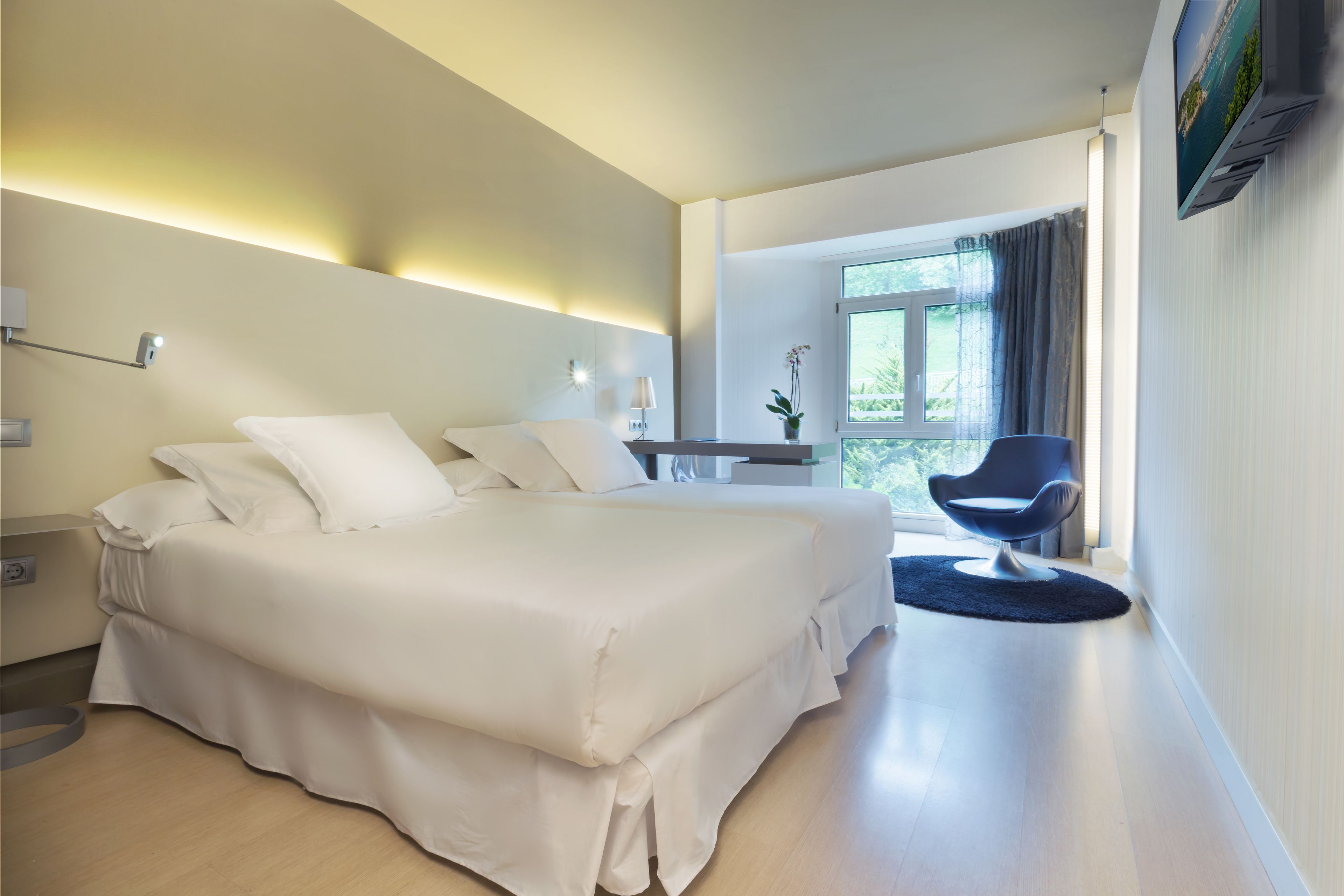 A modern hotel room with a large bed, a chair, a desk, and a window with curtains.
