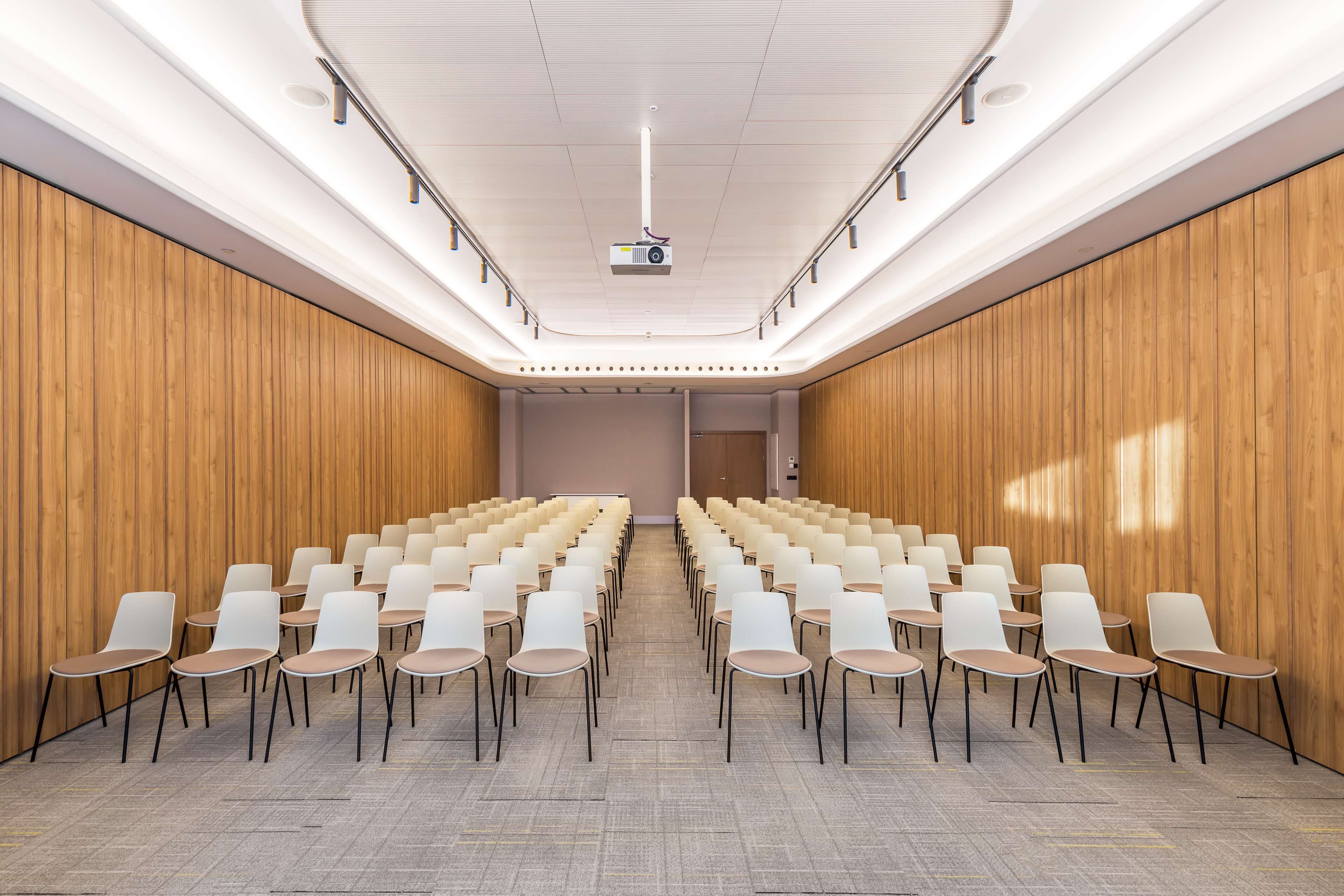 Un salón de conferencias con filas de sillas blancas, un proyector y paneles de madera.