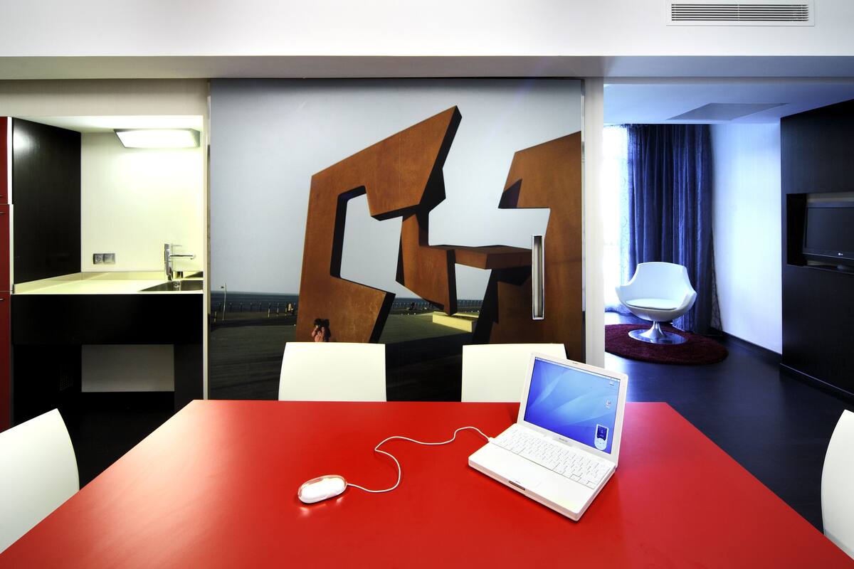 A modern hotel room with a red table, a laptop, a white mouse, and a large abstract wall art piece.