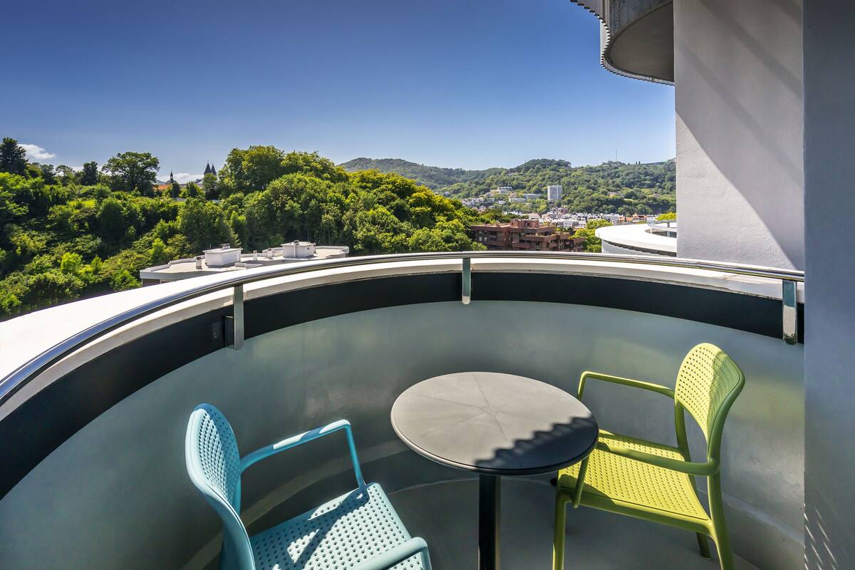 A balcony with two chairs and a table, overlooking a cityscape with greenery and buildings.