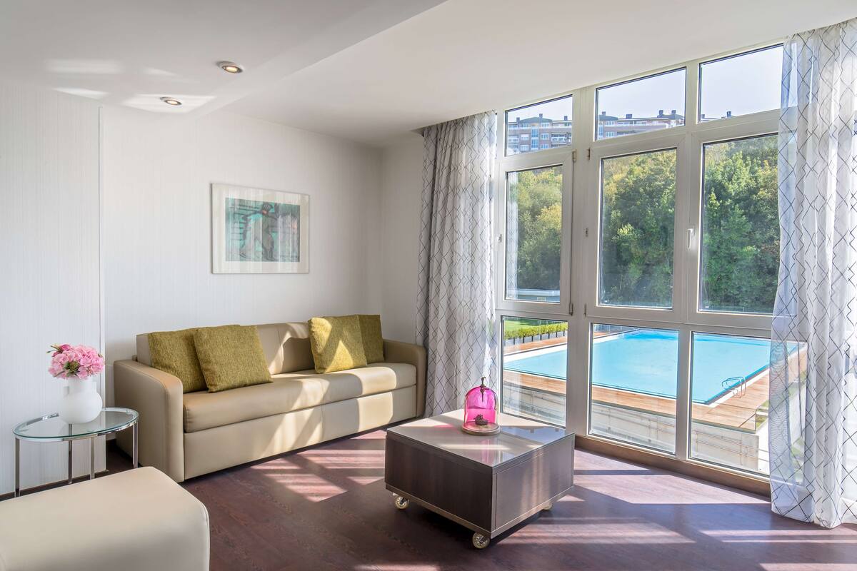 A modern living room with a sofa, a glass coffee table, and a view of a swimming pool through large windows.
