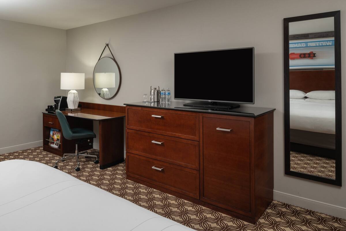 Habitación de hotel con cama, escritorio, silla, televisor, espejo y armario.