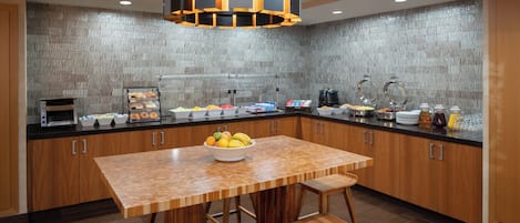 A modern kitchen with a wooden table and bar stools, equipped with a coffee machine, fruit bowl, and various food items.