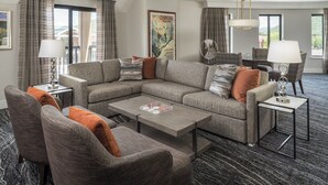 Ein modernes Wohnzimmer mit Ecksofa, Sesseln, Couchtisch und Beistelltisch. An der Wand hängt ein gerahmter Poster, und ein Fenster ist mit Vorhängen drapiert.