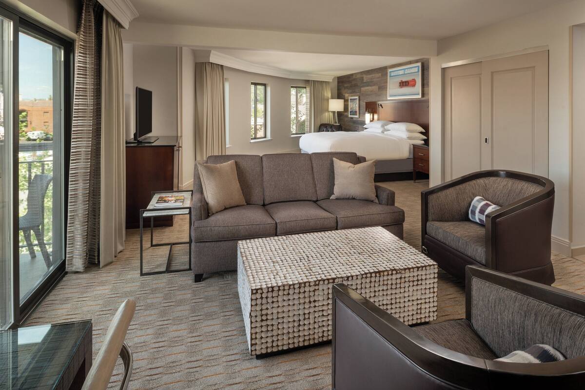 Habitación de hotel con sofá, sillón y una mesita. Se ve una cama al fondo, un televisor sobre un mueble bajo y una ventana con cortinas.