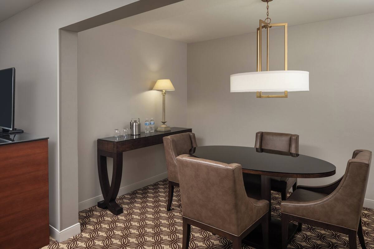 Habitación de hotel con una mesa redonda de comedor, cuatro sillas, un televisor, una lámpara y un minibar.