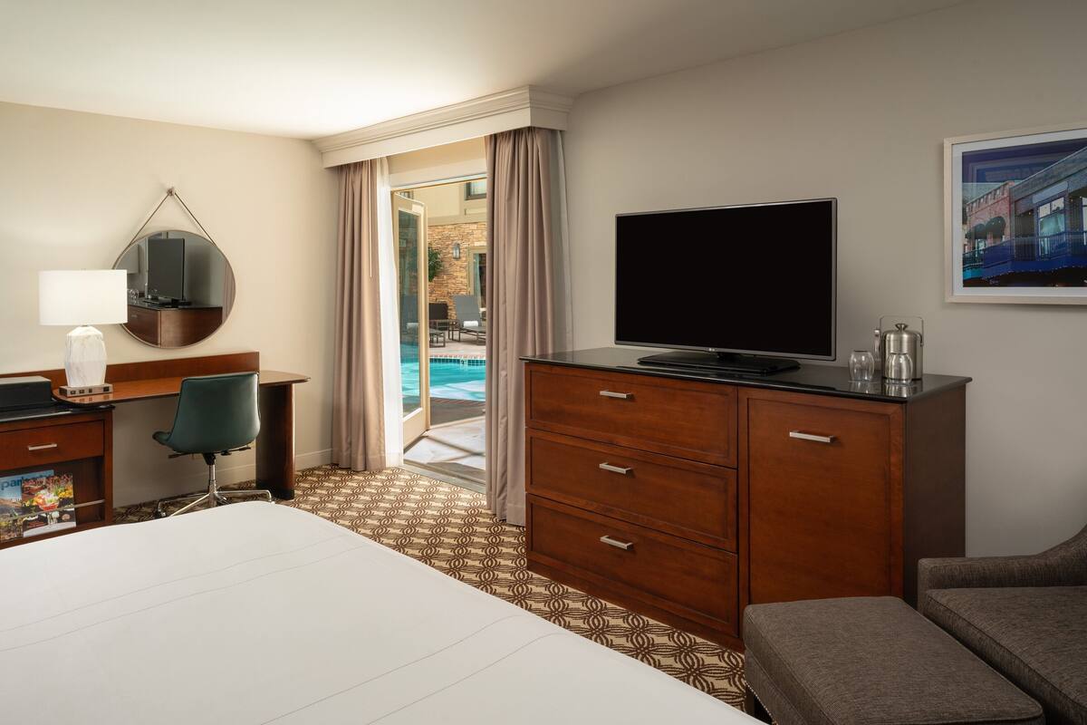 Habitación de hotel con una cama, un escritorio con lámpara, televisión, cómoda y vista a un área de piscina al aire libre.