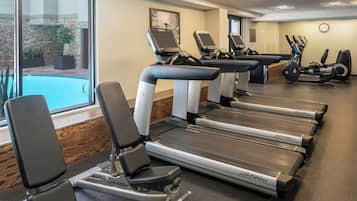 Ein Fitnessstudio mit Laufbändern, Fahrrädern und einem Schwimmbecken, die durch eine Glaswand sichtbar sind.