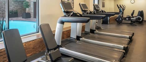 Un gimnasio con cintas de correr, bicicletas estáticas y una piscina visibles a través del ventanal.