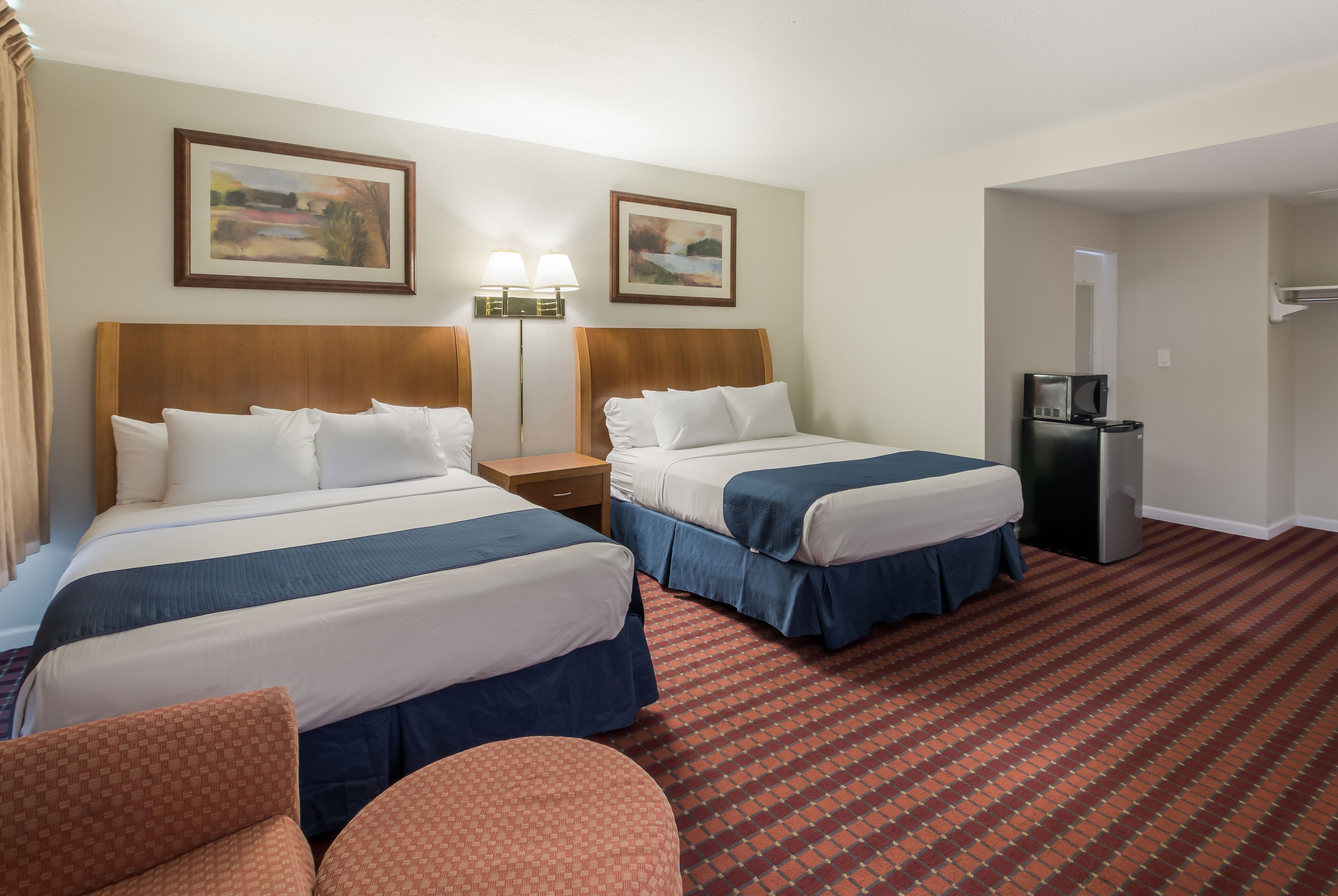 A hotel room with two beds, a desk, a chair, a refrigerator, and two paintings on the wall.