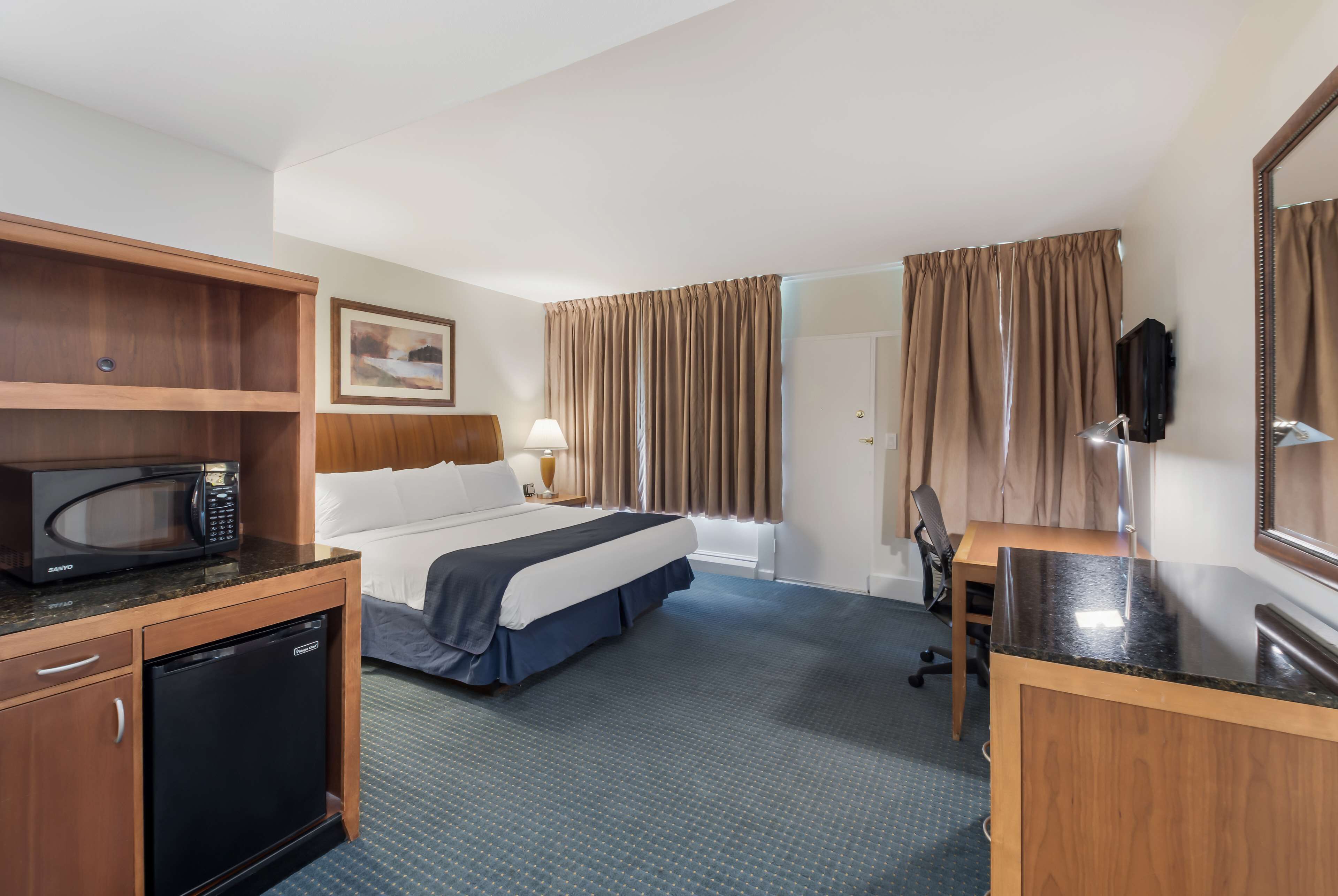 A hotel room with a bed, a desk, a microwave, a refrigerator, a lamp, and a television.