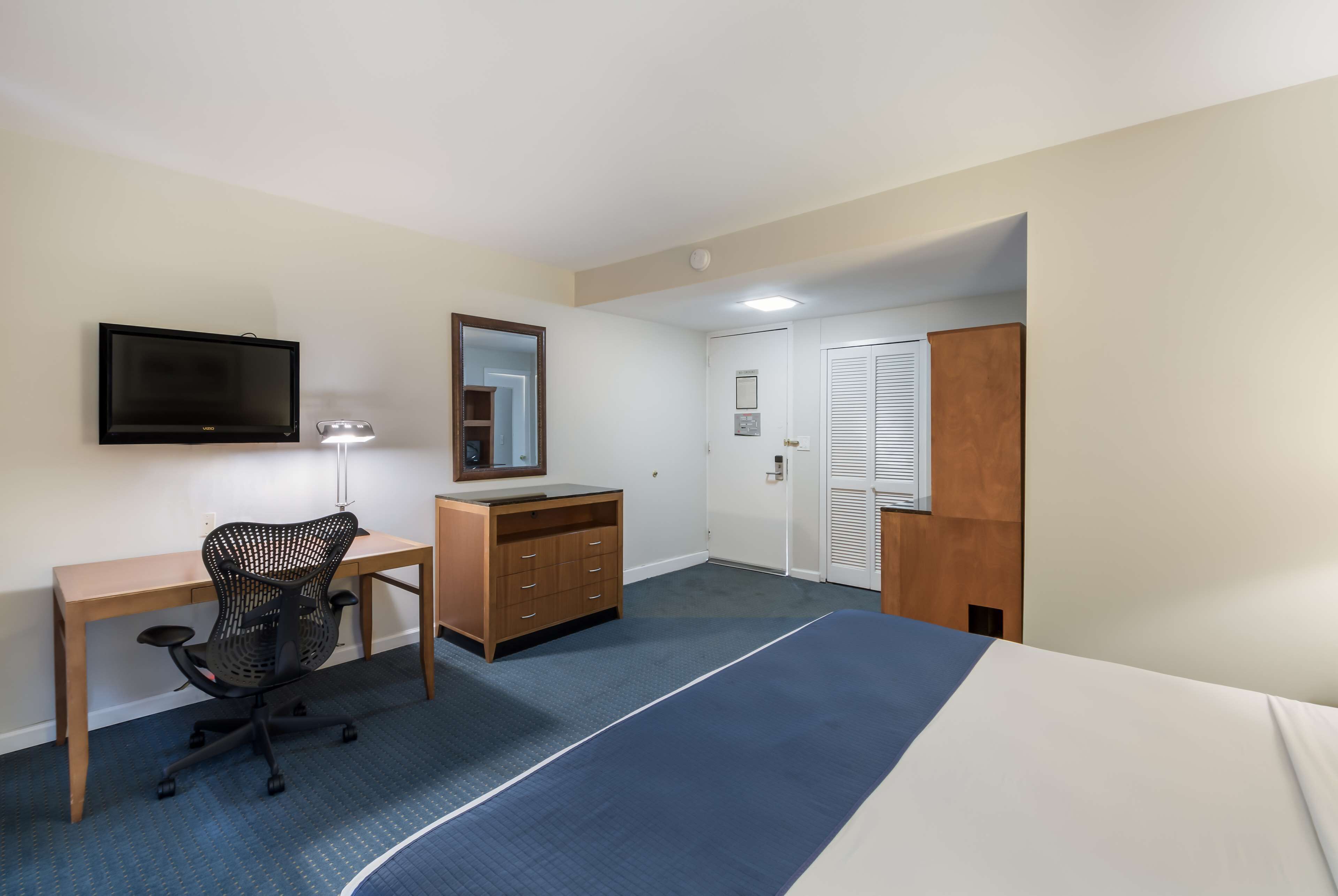 A hotel room with a bed, a desk with a chair, a television, a mirror, and a wardrobe.