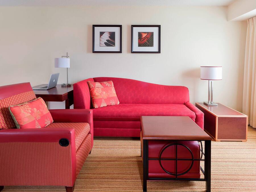 A hotel room with a red sofa, a matching armchair, a small wooden table, a desk with a laptop, and two framed pictures on the wall.