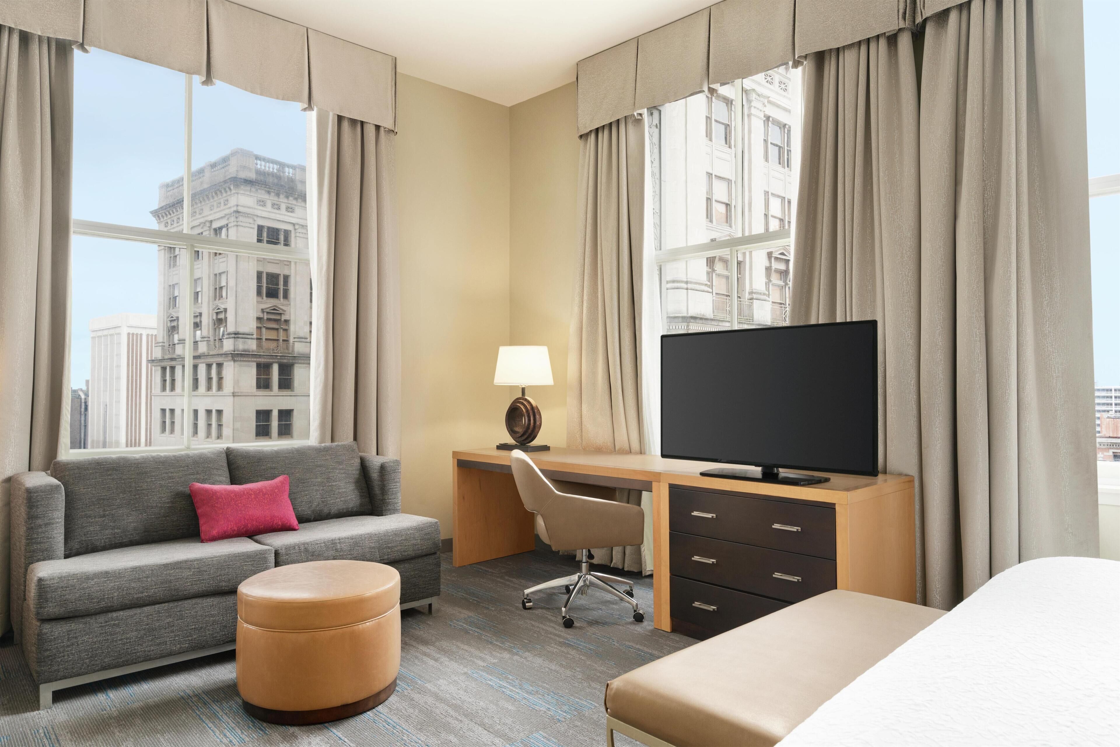 A hotel room with a sofa, ottoman, desk with a TV, and a bed.