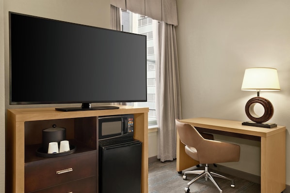 A hotel room with a flat-screen TV, a mini-fridge, a microwave, a desk with a lamp, and a chair.