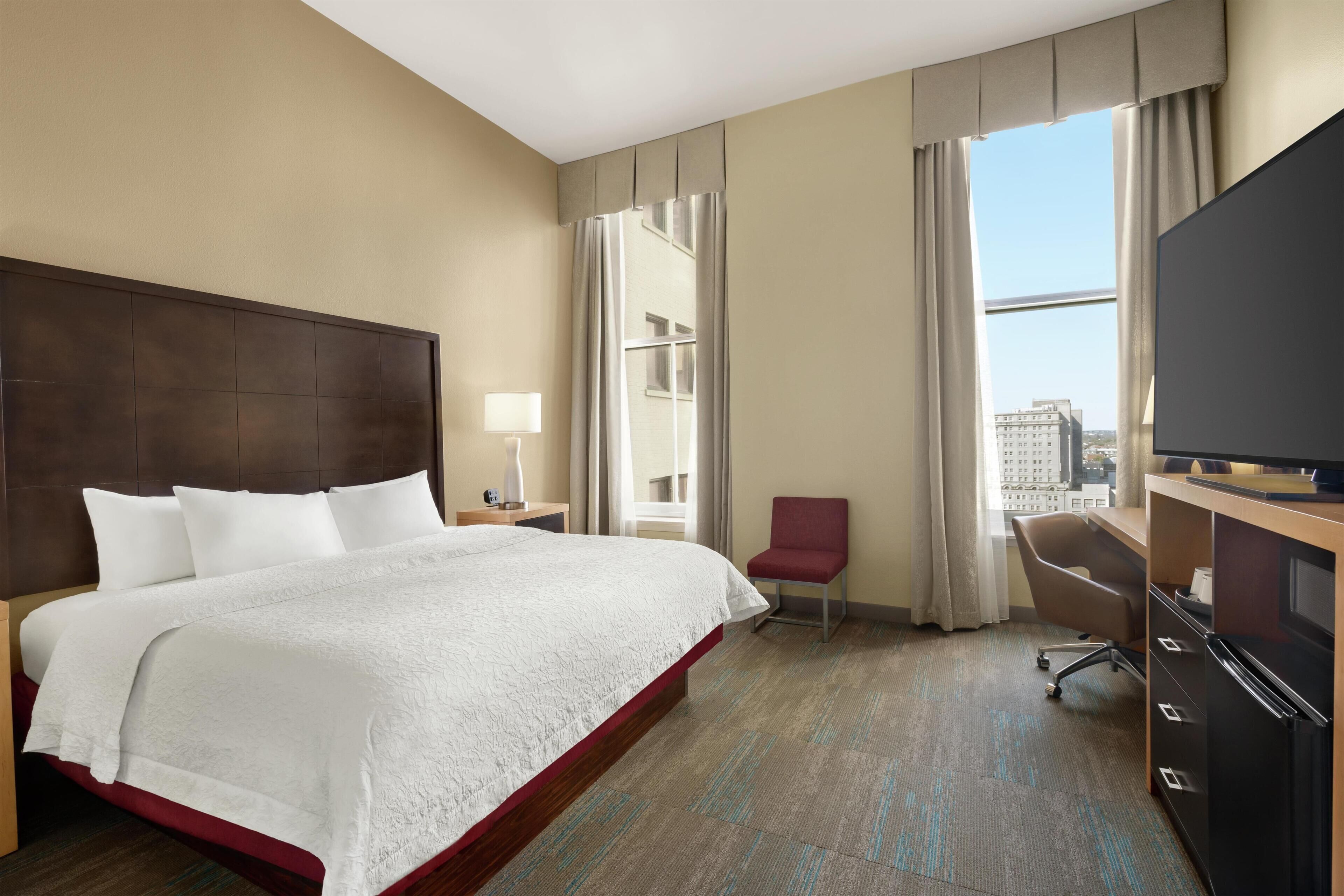 Une chambre d’hôtel avec un lit, un bureau, une chaise, une télévision et une fenêtre avec des rideaux.