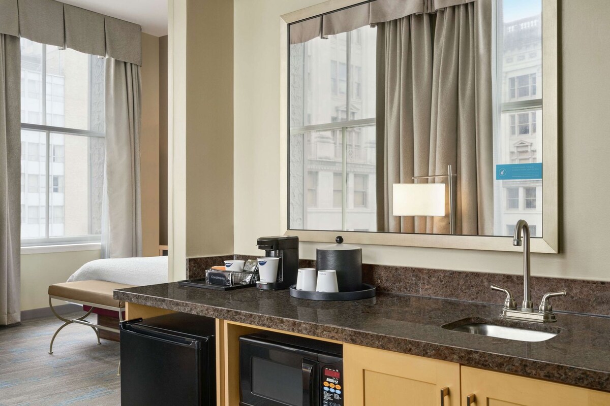 Una habitación de hotel con cama, escritorio con cafetera, lavabo y un espejo grande.