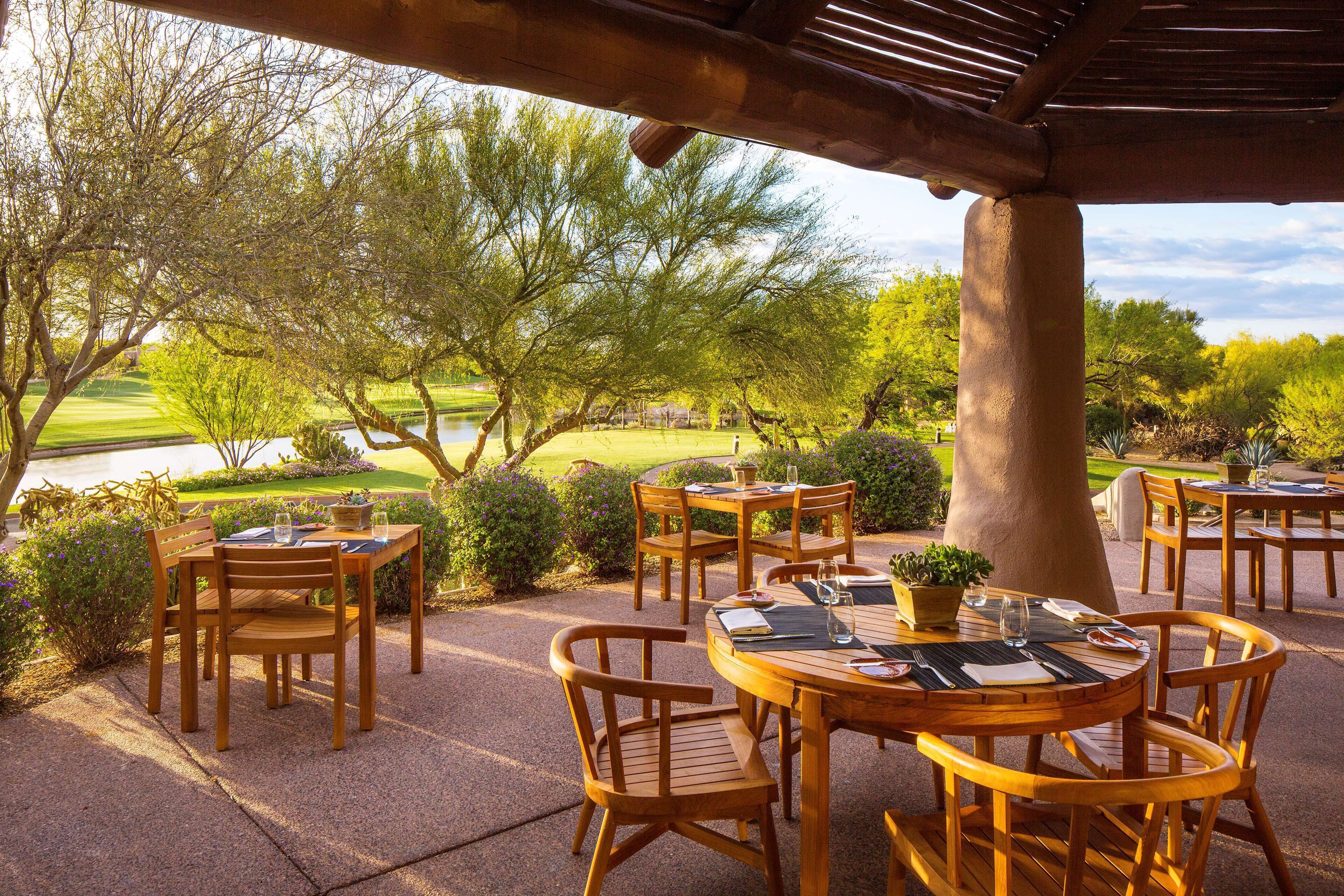 An outdoor patio with wooden tables and chairs, surrounded by greenery and a view of a golf course.