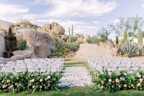 A desert wedding venue with white chairs arranged in rows, a floral aisle, and cacti in the background.