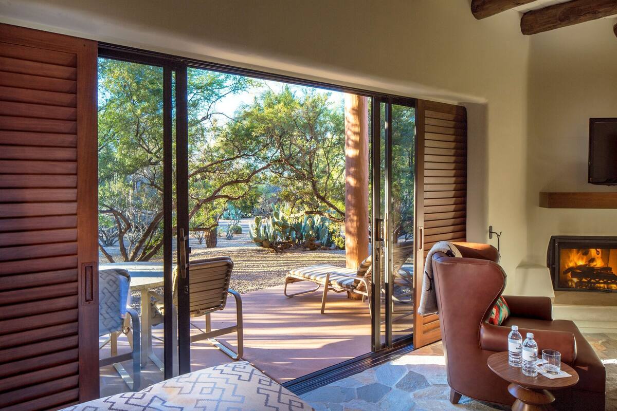 Une pièce avec une cheminée, un canapé, une table avec des bouteilles d’eau et une vue sur un jardin avec des arbres et un cactus.