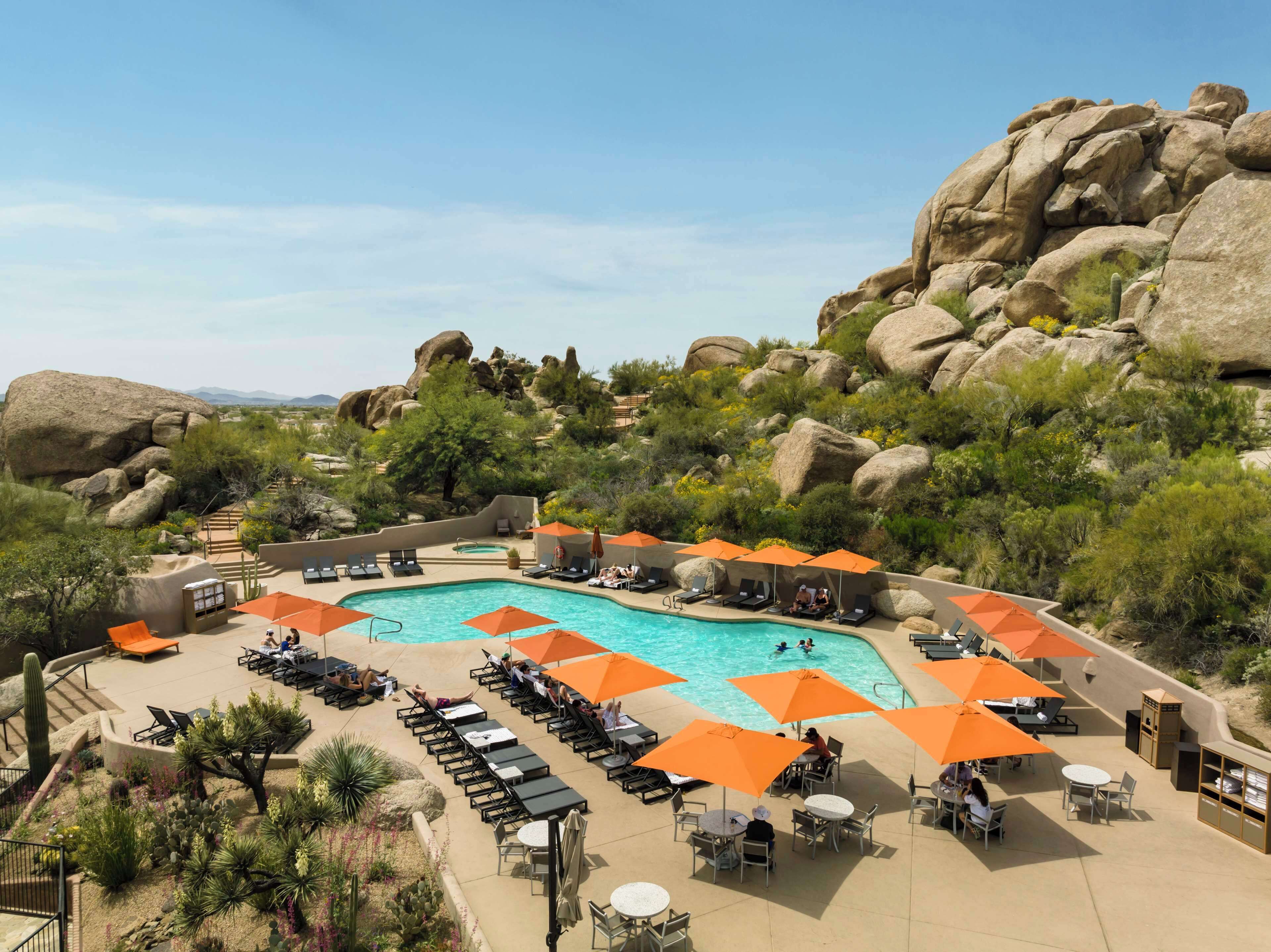 A pool area with lounge chairs and umbrellas, surrounded by large rocks and desert vegetation.