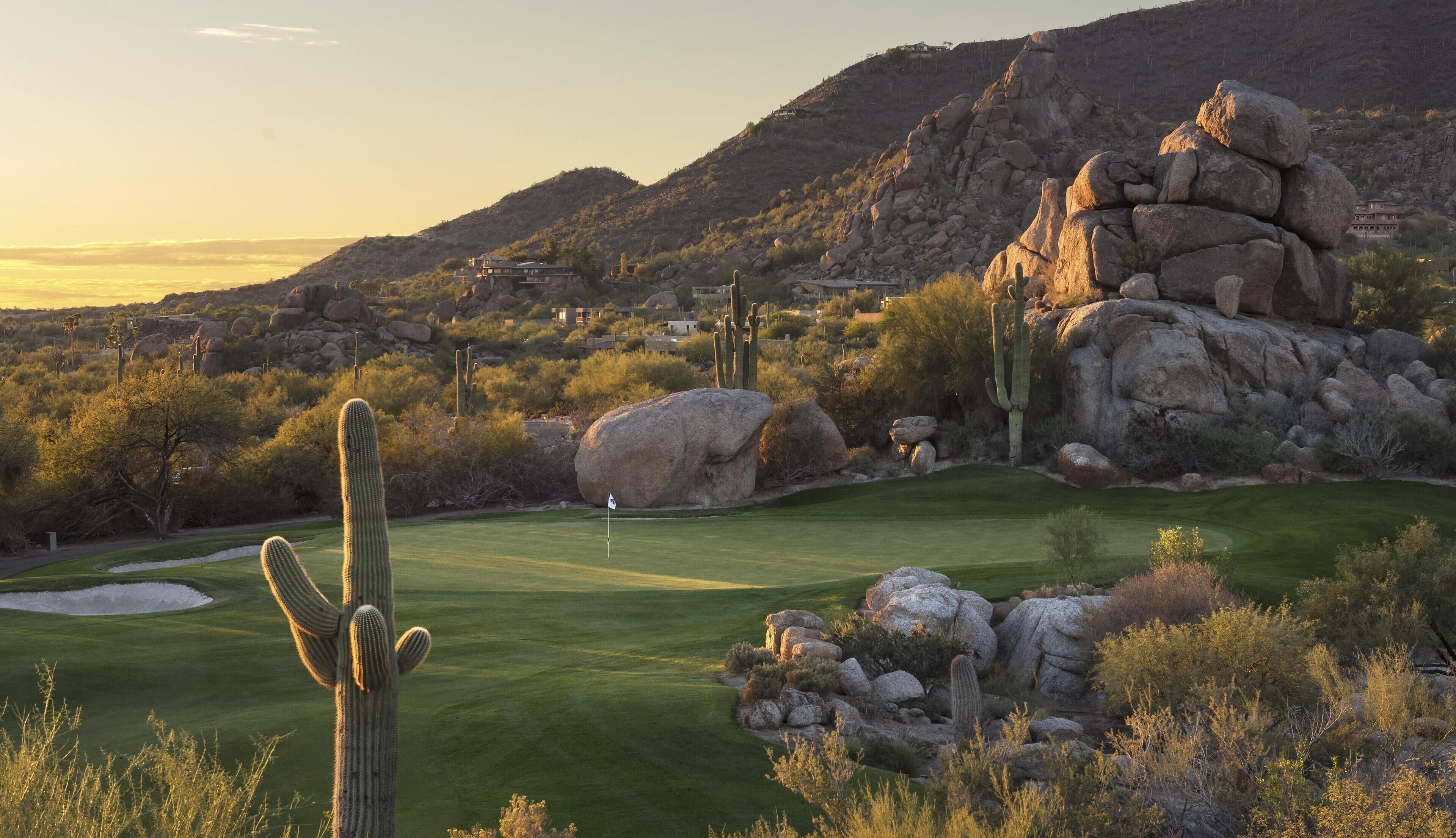 A golf course with cacti and rocks, surrounded by hills and sparse vegetation.