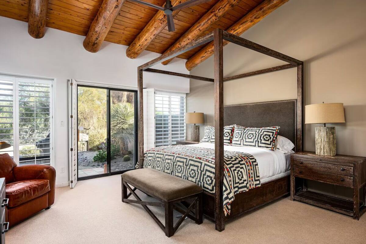 Un lit à baldaquin avec une couvre-lit à motifs, un banc, une table de chevet en bois et un ventilateur de plafond, dans une pièce dotée de grandes fenêtres et offrant une vue sur l’extérieur.