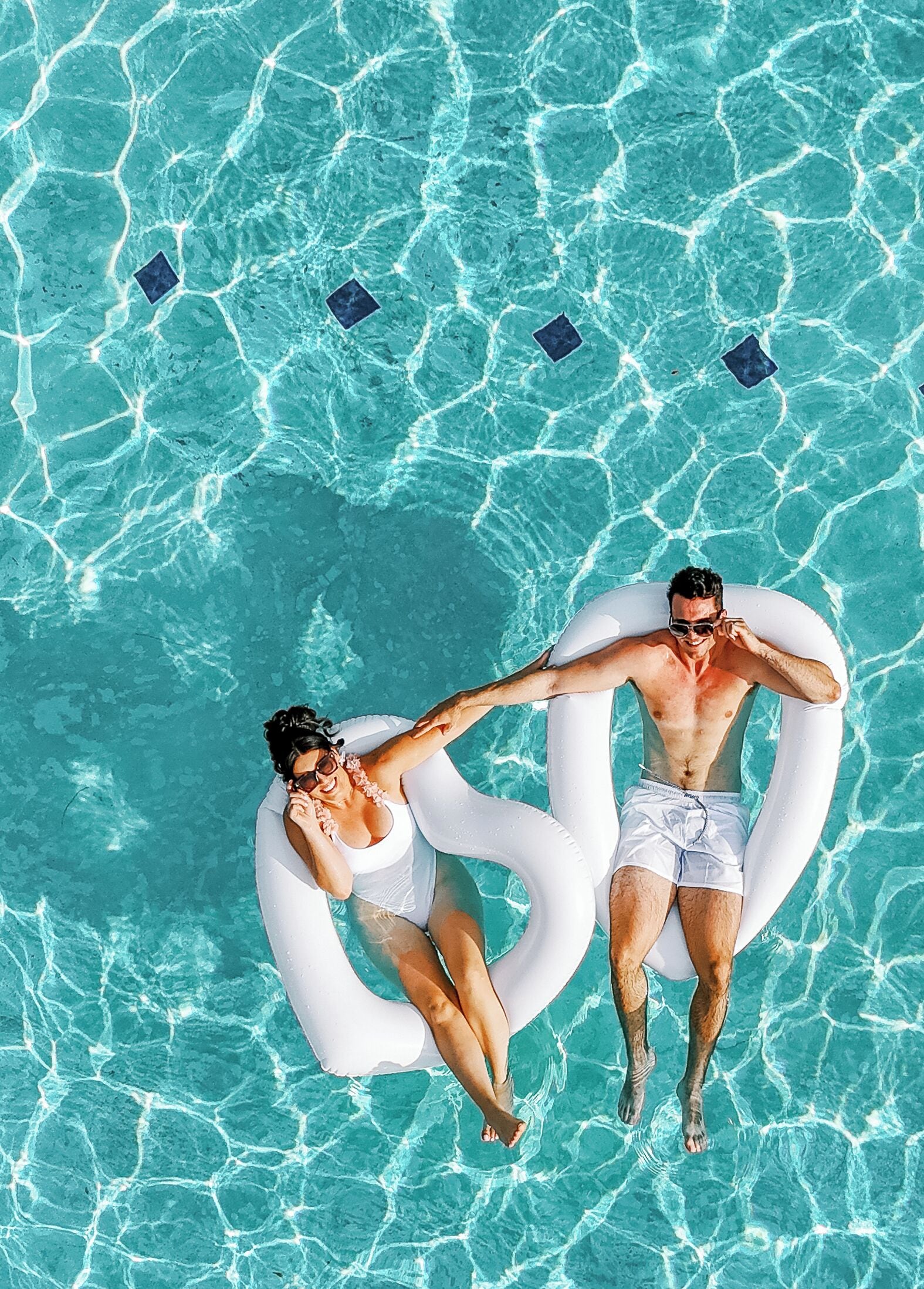 Two people floating in a clear blue pool with white inflatable rings, surrounded by a tiled pool area.