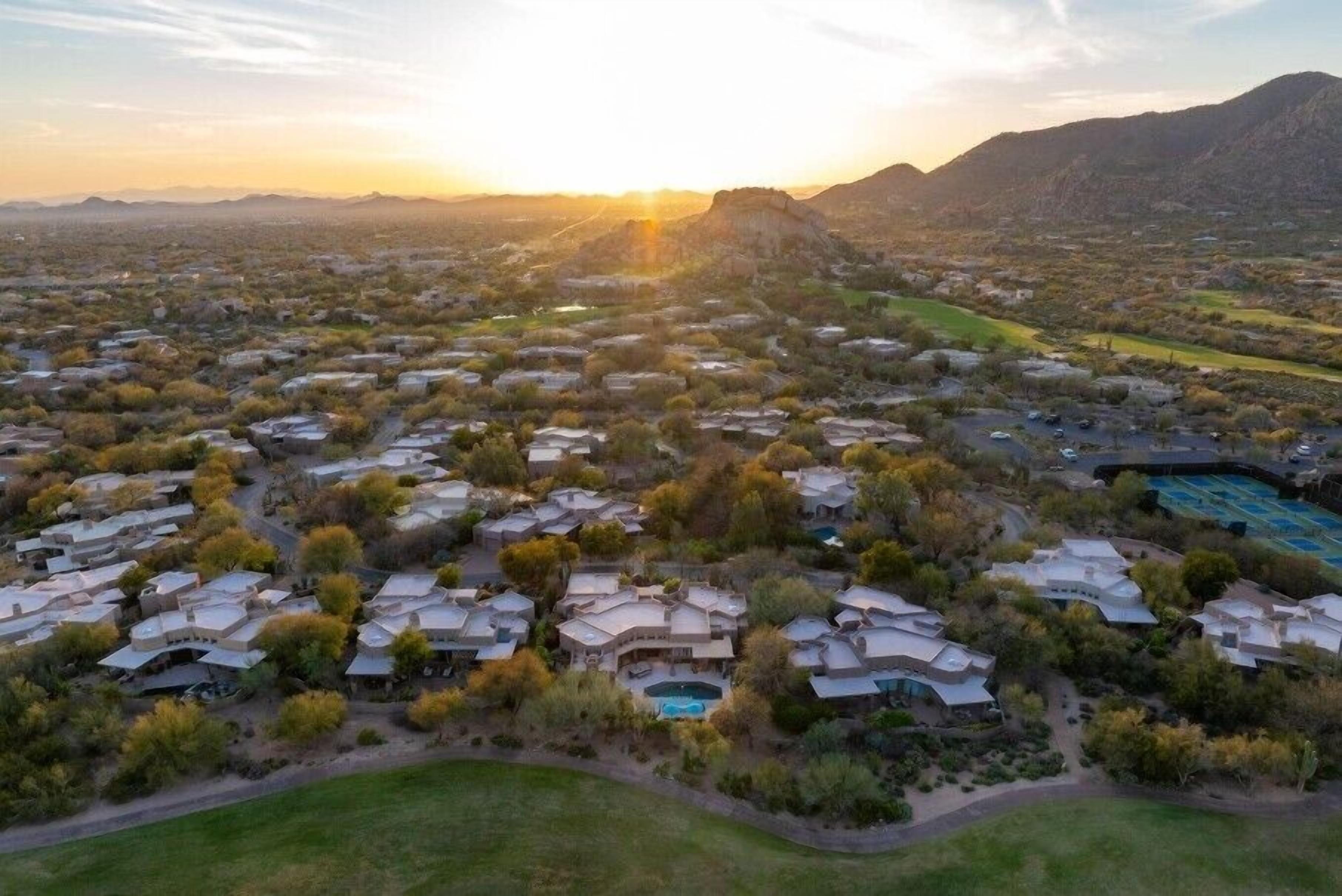 An aerial view of a residential area with multiple houses, a swimming pool, and a hilly landscape in the background.