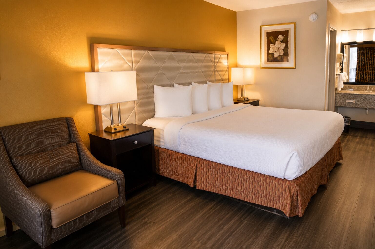 A hotel room with a bed, two bedside lamps, a chair, a nightstand, a framed picture, and a bathroom visible through an open door.