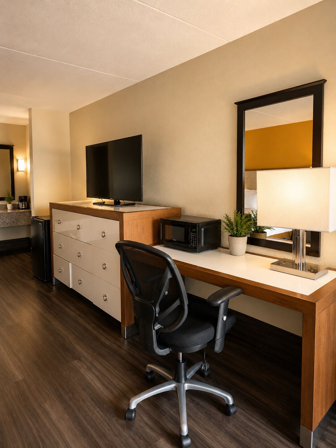 A hotel room with a wooden desk, a black office chair, a flat-screen TV, a microwave, a lamp, and a potted plant.