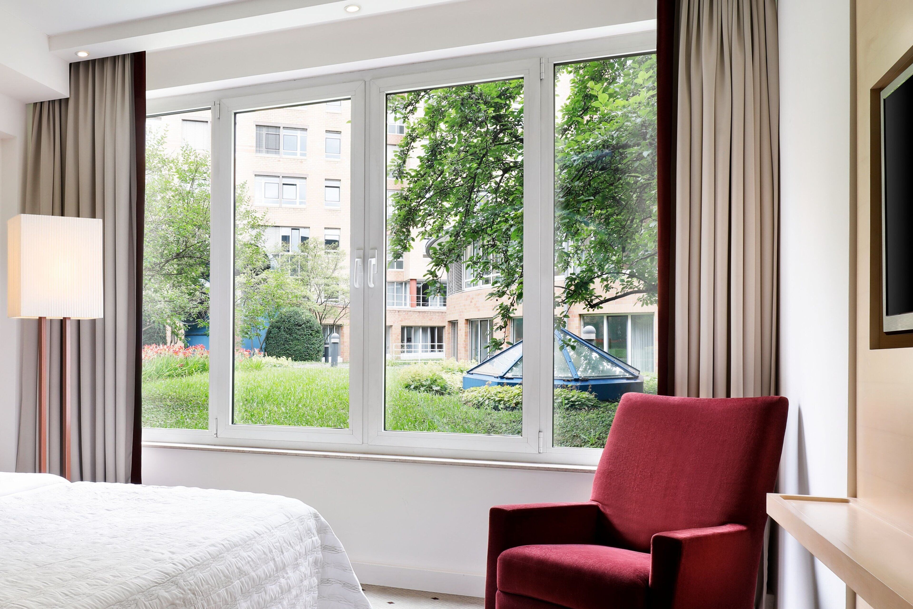 A hotel room with a large window, a bed, a red chair, and a lamp.
