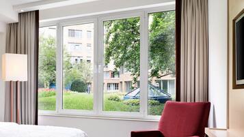 A hotel room with a large window, a bed, a red chair, and a lamp.