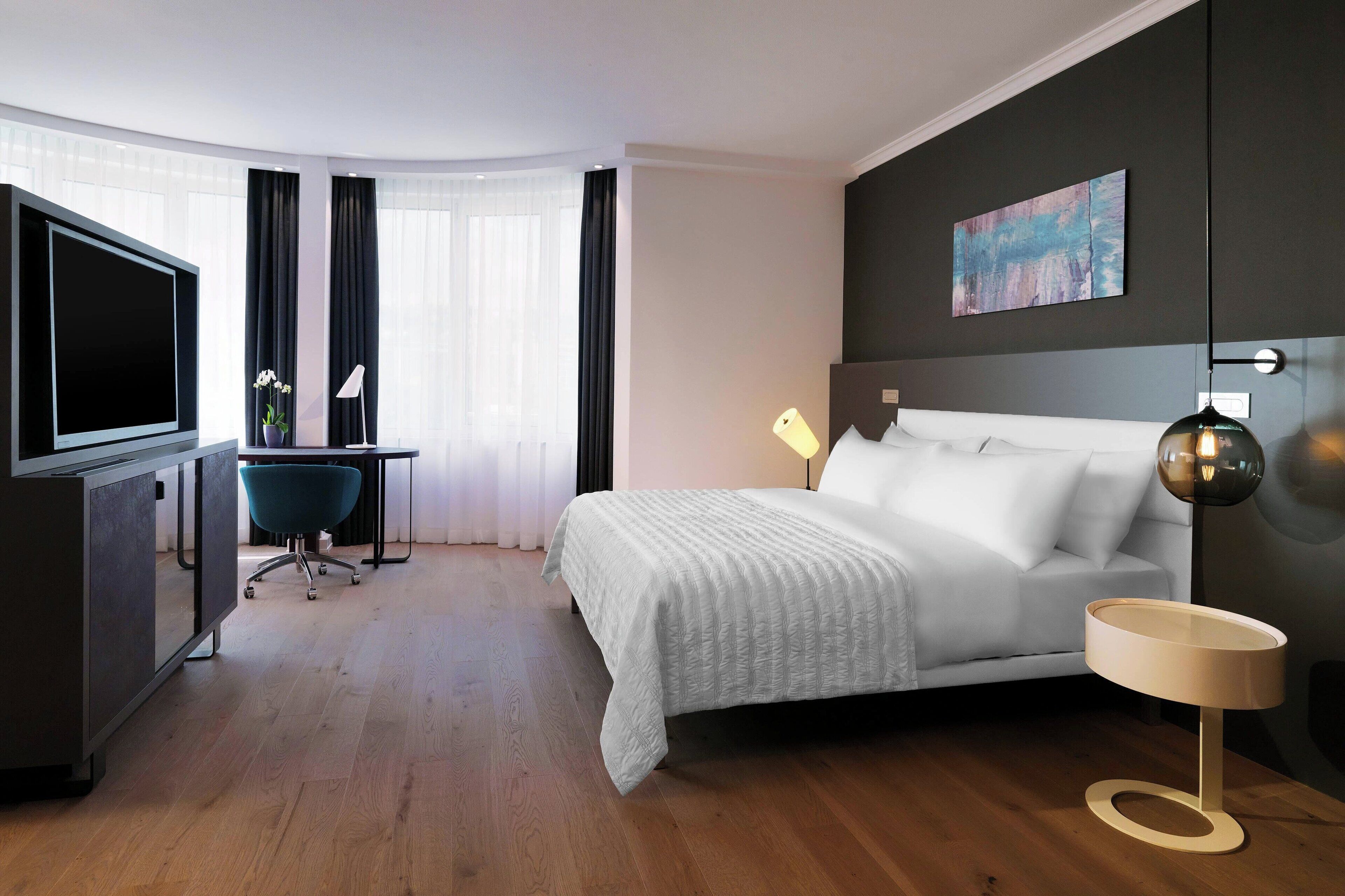 A modern hotel room with a large bed, a desk with a chair, a flat-screen TV, and a painting on the wall.