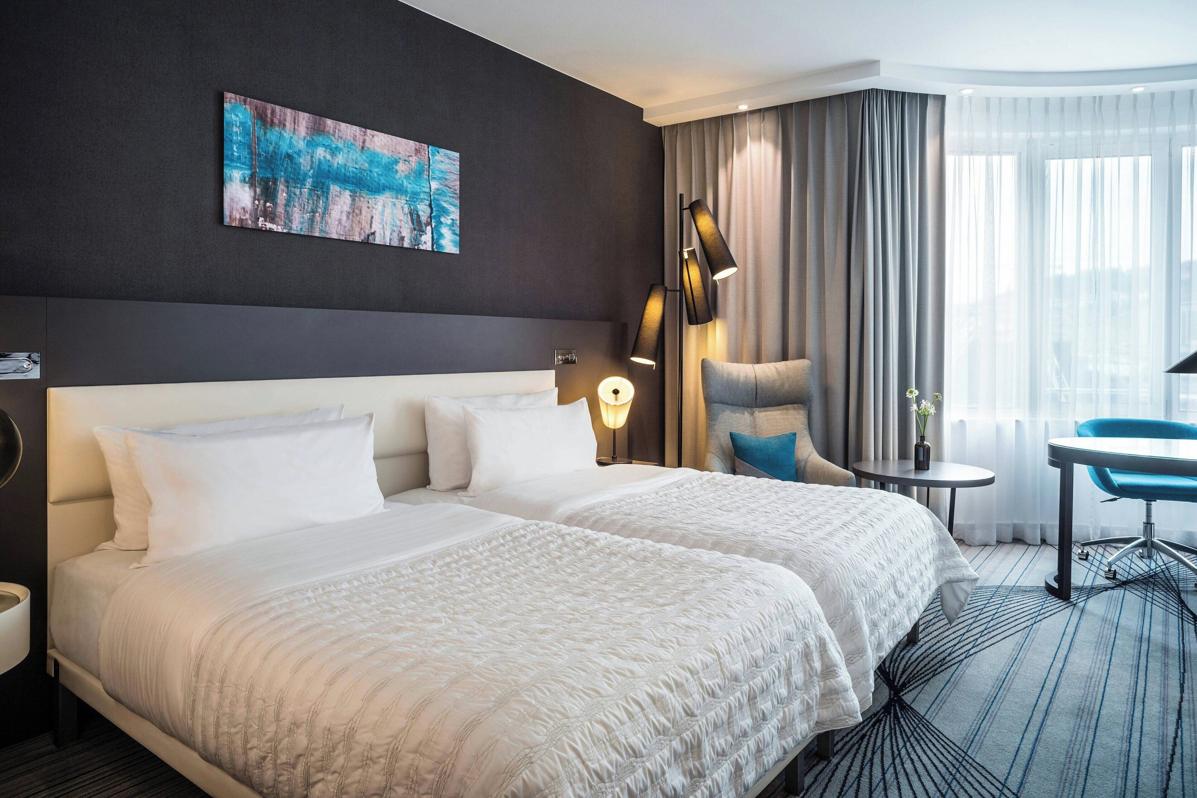 Habitación de hotel con una cama grande, un escritorio, una silla, una lámpara y un cuadro en la pared.