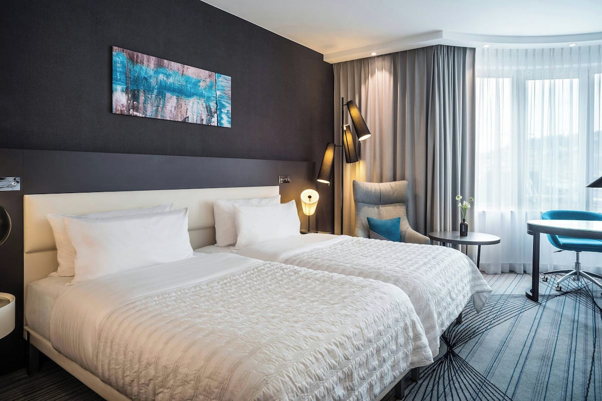 A hotel room with a large bed, a desk, a chair, a lamp, and a painting on the wall.