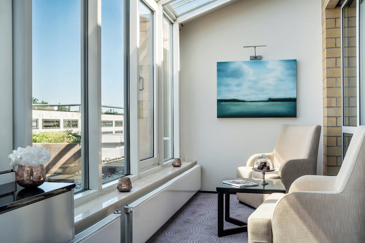 A modern room with a large window, a painting of a landscape, a sofa, and a small table with a vase and a book.
