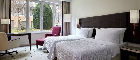 Une chambre d’hôtel avec deux lits, un bureau, une chaise et de grandes fenêtres donnant sur des arbres et des immeubles.