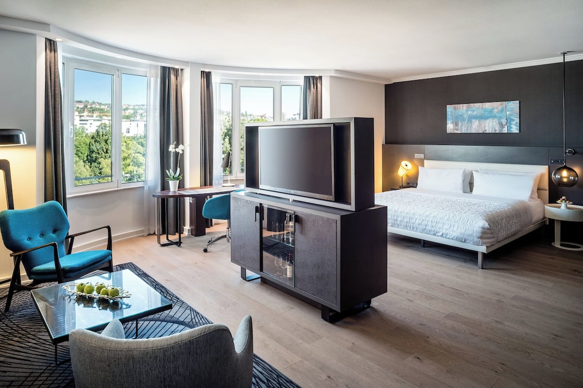 A modern hotel room with a large bed, a TV on a cabinet, a desk with a chair, and a view of the city through large windows.