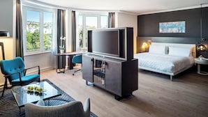 Habitación de hotel moderna con una cama grande, un televisor en un mueble, un escritorio con una silla y vistas a la ciudad a través de amplias ventanas.