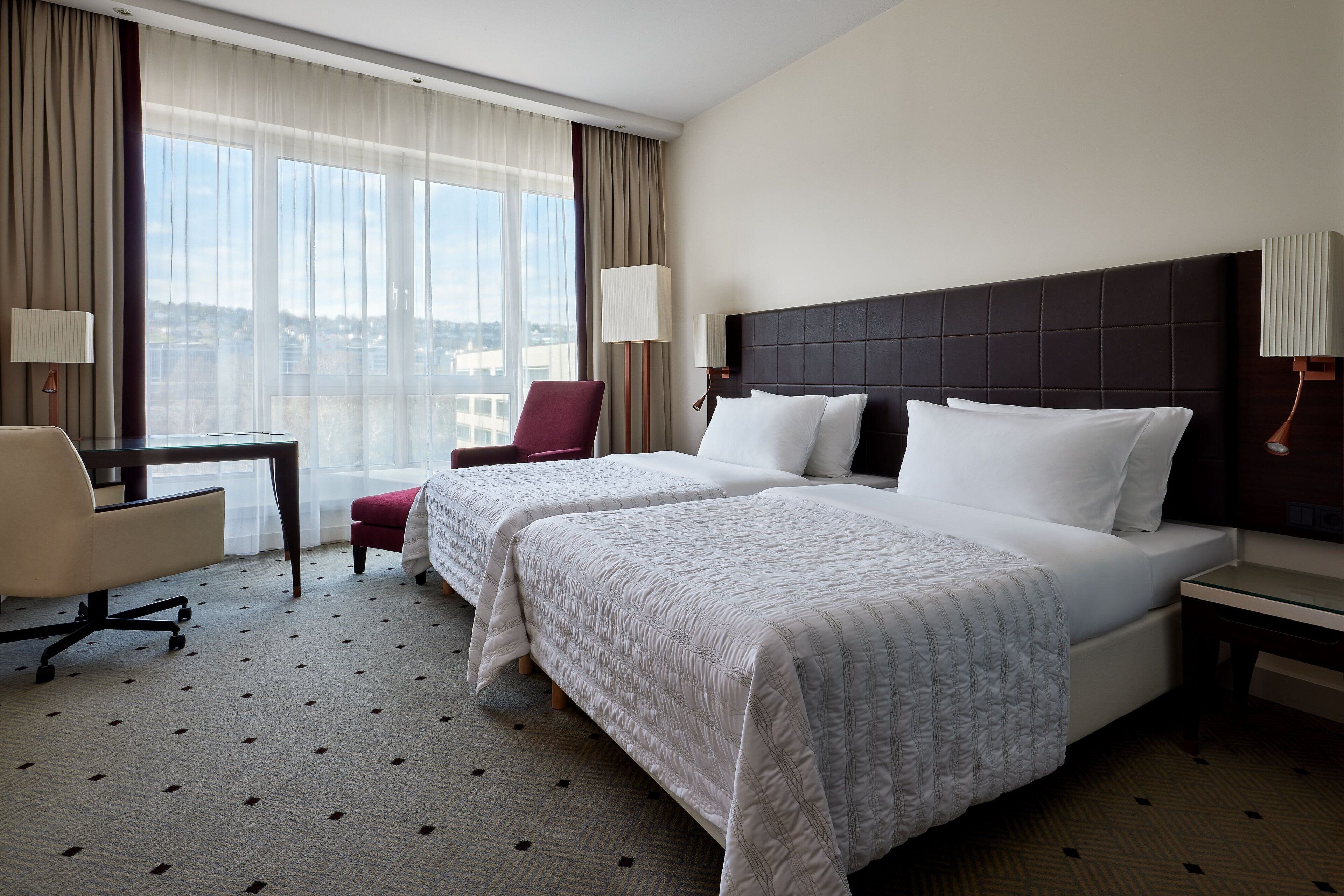 Une chambre d’hôtel avec deux lits, un bureau, une chaise et une grande fenêtre donnant sur la ville.