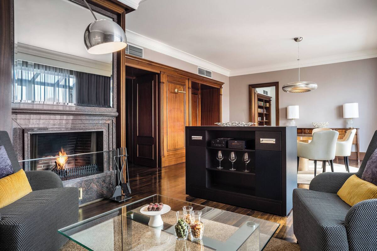 A modern living room with a fireplace, dark wood furniture, a glass coffee table, and a dining area with white chairs.